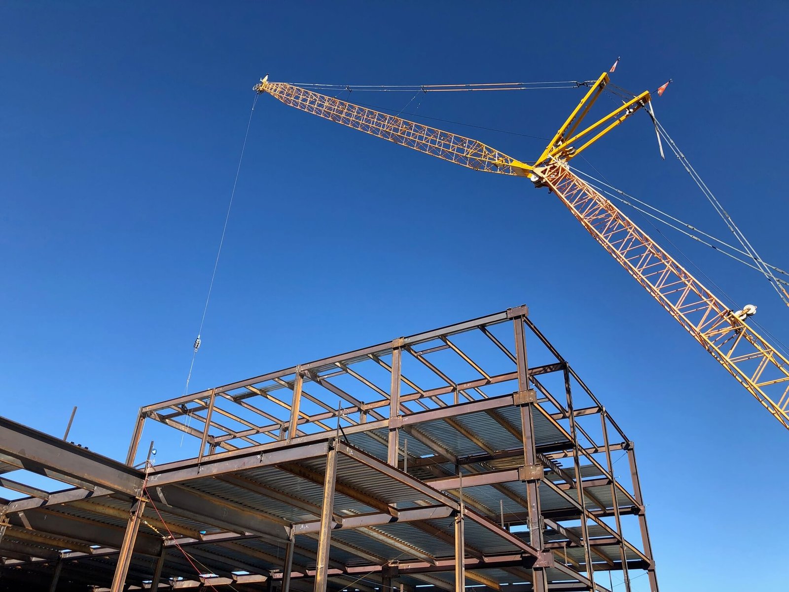 A large crane lifting steel beams during the erection of a multi-story structural steel framework under a clear blue sky.